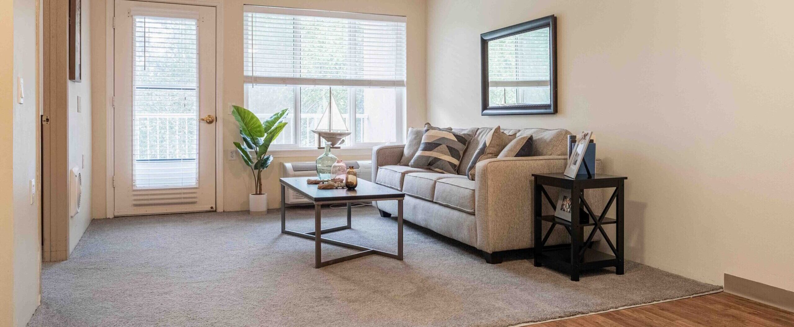 A living room with a gray couch with throw pillows on it, a coffee table with three vases, a side table with photo frames on it, and a potted plant by the door leading to the balcony at Augustine Landing near Orange Park, FL.