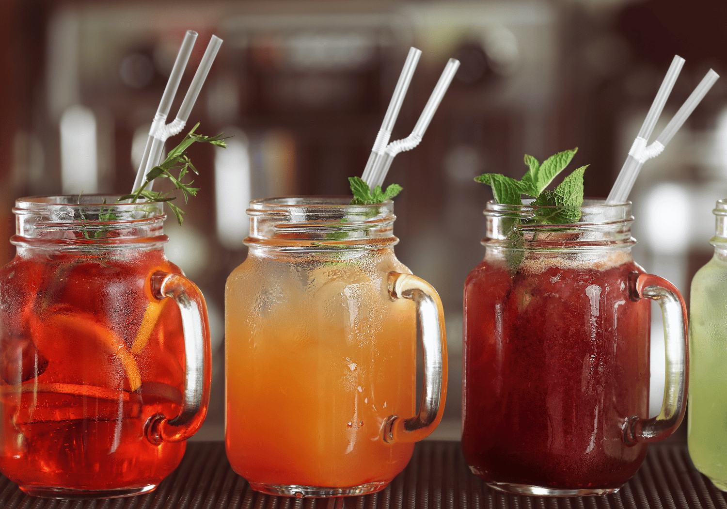 Three colorful mason jar drinks with ice, mint garnish, and white straws on a bar counter.