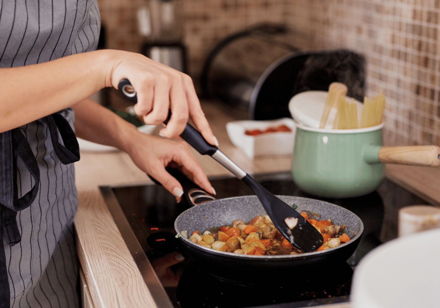 Hands in a striped apron stir-fry diced vegetables in a speckled frying pan on a stovetop, using a black spatula.