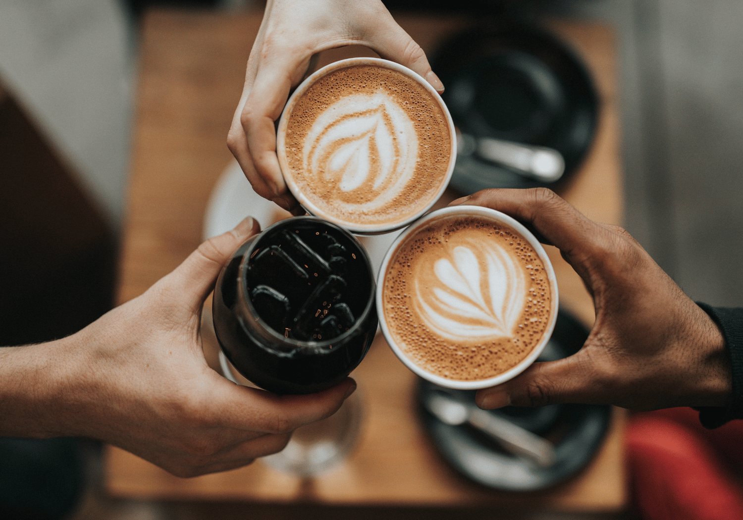 Three hands hold coffee cups with latte art as they toast, with a glass of iced drink on a wooden table nearby.